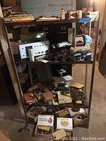 Metal shelving unit displaying various items including radio amplifier, boxes, cables and components