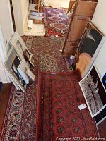 Three oriental style area rugs laid out in a room with various framed artworks leaning against walls. Rugs show intricate floral and geometric patterns in red, blue, and beige, appear worn and need cleaning.