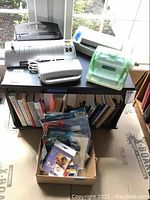 Wide view of three laminators on a black table with assorted laminating supplies in a box beneath it, including sheets and books on shelf below.