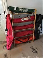 Seven camp chairs in various colored carry bags resting on a wooden rack; rack not included in sale.