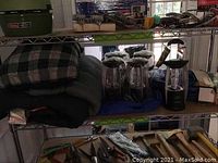 Shelving unit with folded Coleman plaid sleeping bag and green insulated sleeping bag, and five Remington battery powered lanterns in a row on shelf below.