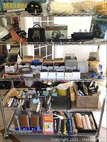 Overview of metal shelving loaded with measuring wheel, gloves, tool belt, discs, painting and sanding supplies