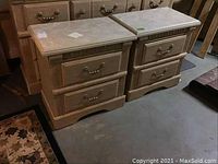 Pair of matching wooden nightstands with faux marble tops, two drawers each, decorative handles and carved accents, placed side by side on a concrete floor.
