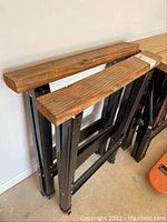 Full view of two foldable sawhorses with wooden tops and black metal foldable frames, positioned upright and leaning against a wall.