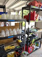 Two plastic shelving units in garage with items on top and shelves. One shelf is black with black legs. Another shelf is black with gray legs. Items on shelves include glass lamp shades and tools but are excluded from sale.