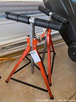 Single board stand showing orange tripod legs, black rubber feet, adjustable height stem, and black roller bar on top