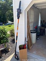 Remington extendable pole saw standing against a garage exterior wall with a cardboard box containing loppers and pruning shears inside the garage door opening.