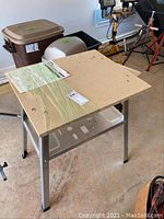 Workbench placed in garage showing metal legs and rectangular wood composite top with assembly instructions under plastic wrap.