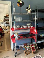 Full view of the metal shelving unit with the 5 vented shelves on caster wheels showing various glassware, china, and some decorative items.