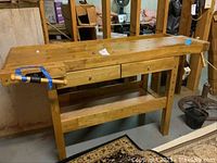 Front view of solid wood workbench showing full-length top, drawers, vise at front left, and lower shelf.