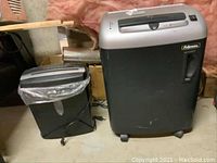 Photo of two paper shredders side by side in basement storage, showing size difference and general appearance.