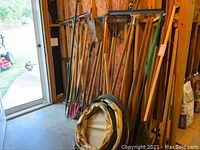 Wide view of the tools leaning against a wooden framed wall near a door, includes shovels and other long-handled tools.