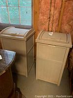 Pair of beige Suncast outdoor trash cans positioned side by side inside a basement with exposed insulation, showing full front and partial side views.