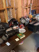 Sports equipment displayed on a wooden table in an unfinished basement showing baseball catcher's protective gear including Riddell helmet and Easton leg guards, along with football shoulder pads, a frisbee, and some boxed board games.