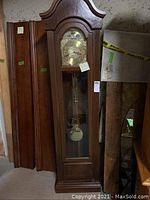 Full view of the grandfather clock standing upright showing the wood casing, arched top, glass door with pendulum inside.