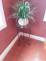 Artificial faux green grass plant with fuzzy white flower spikes in woven basket pot placed on a black metal plant stand with curved legs, next to red wall and tiled floor.