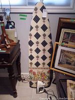 Full view of ironing board with patterned cover leaning against a wall next to framed art and a table; Black & Decker iron resting on floor in front.