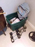 Multiple dumbbells placed on floor next to green plastic bin containing additional exercise items and a backpack bag.
