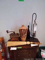 Photo of a collection including a vintage coffee grinder with wooden base and drawer, a rustic bark vase, black bottle with cap, woven broom, and an iron hanging candle holder with glass enclosure.