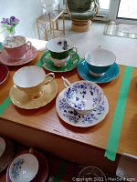Four assorted vintage teacups and saucers displayed on wooden table with green painter's tape, showing different designs and colors including gold, green, pink, and blue/white floral patterns.