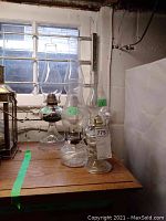 Five glass oil lamps on a wooden surface near a window with natural lighting showing details of chimneys and reservoirs.