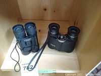 Two black binoculars on a wooden shelf, one Tasco 571 and one Bushmaster model