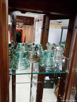 Five glass electrical insulators displayed on a glass shelf inside a wooden cabinet, showcasing shapes and green and clear glass coloration.
