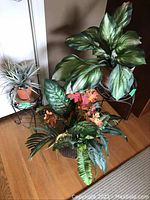 Three faux plants in terracotta pots on and near black metal plant stands, showing details of leaves and flower accents.
