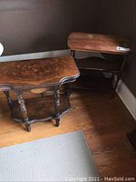 Two vintage wood veneer side tables positioned on wooden floor with rug; one features a magazine/book rack below top surface, displaying carved legs and dark finish.