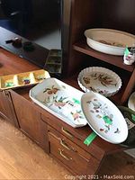 Overview of three Royal Worcester dishes on wooden furniture: a small pot with lid, a fluted round dish, and an oval platter with fruit designs.