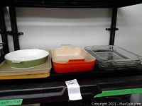 Photo showing all five baking pans on metal shelving: Pampered Chef beige baking sheet on the bottom, CorningWare round dish, Le Creuset orange lasagna pan, and two clear glass lasagna pans side by side.