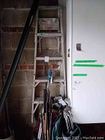 Photo of a gray aluminum step ladder with paint spots and wear marks leaning against a brick wall with various tools and golf clubs below it.