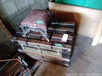 Wooden steamer trunk with brass hardware and black metal bands topped by cast metal stool with worn upholstered top, both showing wear and rust.
