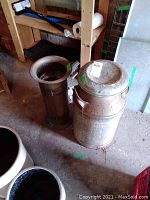 Photo showing metal milk can and brass umbrella stand in a garage setting with visible wear and rust on milk can and patina on umbrella stand