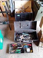 Three toolboxes arranged in a garage setting with the wooden toolbox at the back and two plastic toolboxes open showing assorted hand tools and electrical cords inside.