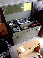Green metal tool box open showing tools inside on lift-out tray, including drill bits set, screwdriver, saw, and other tools.