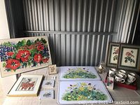 Photo showing assortment of framed artwork, two floral placemats, ceramic teapot, four matching mugs, and small floral prints on a table.