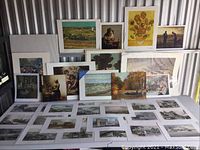 Photo showing entire lot of assorted framed and unframed prints arranged on and behind a table in a storage unit.