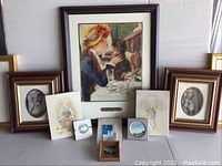 Front view of collection showing a large framed print of a woman with a cat, two oval framed children's portraits, and smaller unframed illustrations and cards arranged in front.