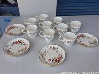 Photo showing 7 matching teacups and 6 matching small dessert plates with floral pattern and gold trim, arranged on a grey surface.