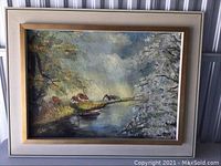Framed landscape painting showing river, houses, and trees.