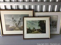 Two framed art prints leaning against metal wall, showing muted landscape scenes with trees and rural houses.