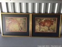 Two framed art prints featuring prehistoric animal depictions side by side, showing the gold frames and black/red mats under light with some reflections and dust visible.