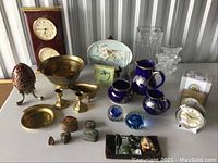 Full lot on table showing all items described including wooden clock, brass chalice, candlesticks, blue pitchers, decorative plate, glass vase and paperweights, and assorted small figurines and trinkets.