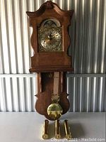 Front view of wooden pendulum wall clock with brass pendulum and weights removed and placed below the clock case.