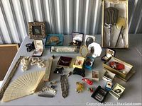 Overview of entire lot on table: framed art, necklaces, fan, vanity set, watches, jewelry boxes, brooches, rings, compact set