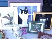 Photo showing multiple framed artworks including large cat painting, framed bird prints, and small garden scene print, along with some ceramic items on table.