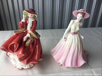 Front view of two porcelain ladies figurines, one in red coat and hat, the other in pink and white dress with pink hat.