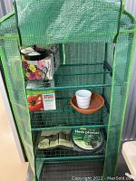 Front view of 60 inch portable greenhouse showing four metal wire shelves, green mesh covering, partially open front flap, and various gardening items on shelves (not included in sale).