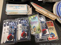 Overview photo showing candle powered boat in original box, wooden sailboat model, and multiple smartphone pet ID tags in packaging with Star Wars, Disney Fairies, and Mickey Mouse designs.
